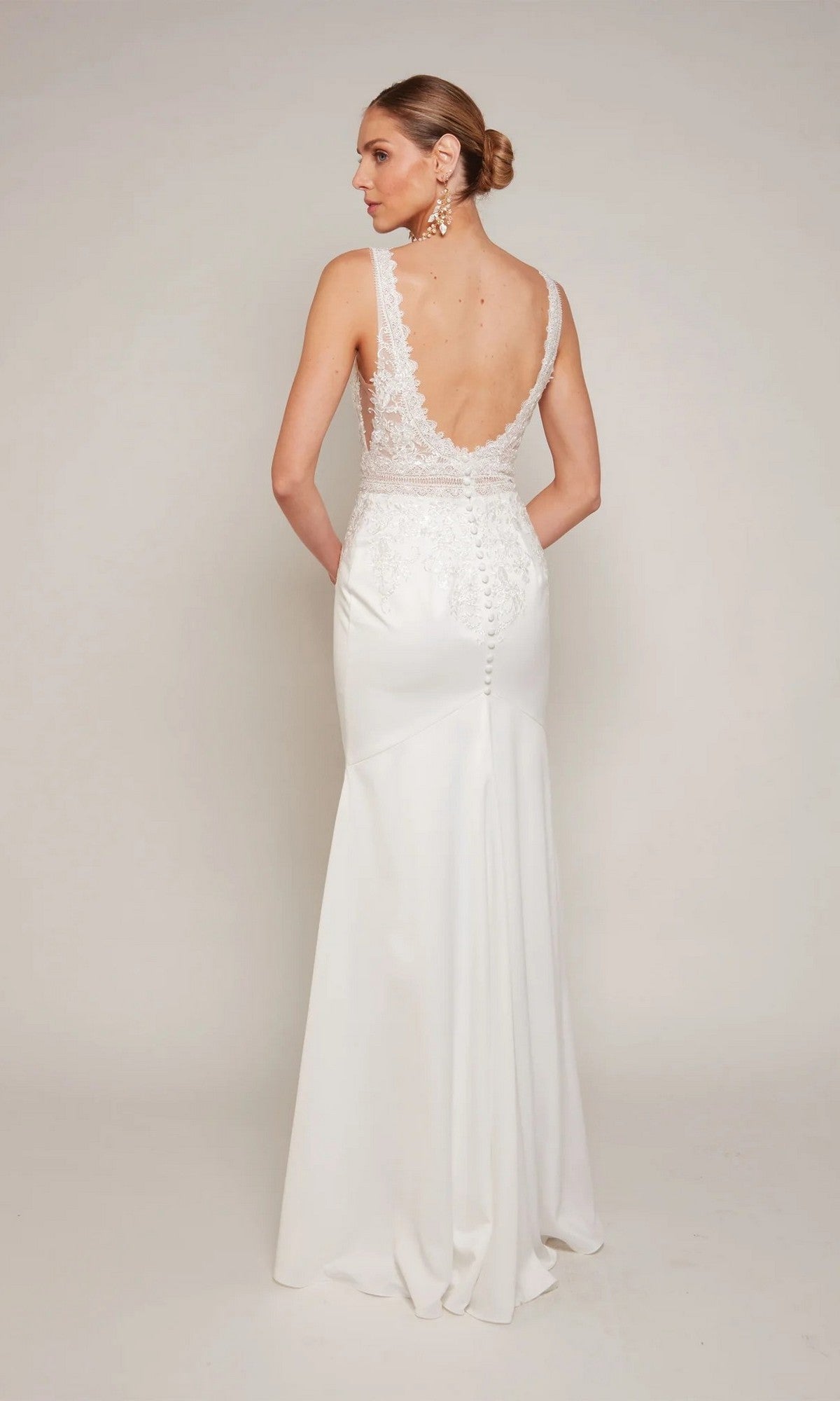 A woman stands with hands behind her back, facing away in the Alyce 7097 Long Formal Dress—a sleeveless gown featuring lace appliqué and a low back, set against a plain background.